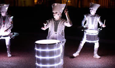 Figures elaborately dressed and covered in lights play the drums