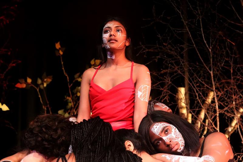 medea actress of colour in a red top, surrounded by the chorus wearing black clothes. All have tribal patterns on left hand of face in white