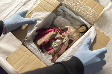 Two hands wearing blue nitrile gloves open a carton box which contains a doll. The doll is brunette and wears a red and beige dress and a beige lace shawl.