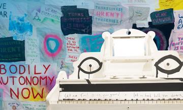 White piano decorated with black painted eyes in front of blue wall with posters, one reading 'BODILY AUTONOMY NOW'
