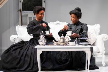Two black women sit on a white chaise lounge drinking tea