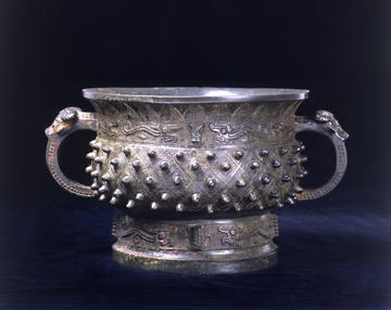 A bronze food basin, standing on a high foot with two handles, with bosses within diamond patterns. Just below the neck, around the body, is a band displaying dragons, and similar dragons decorate the foot.