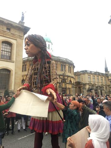 A large puppet of a young girl stood in the middle of Broad Street Oxford, surrounded by people and holding a piece of paper.
