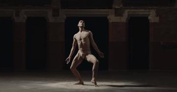 Image is a still photo from 'Jon's solo' dance performance depicting a dancer in the spotlight of an otherwise dark room. The body is in a pose as if attached to threads like a marionette. 