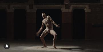 Image is a still photo from 'Jon's solo' dance performance depicting a dancer in the spotlight of an otherwise dark room. The body is in a pose as if attached to threads like a marionette. 