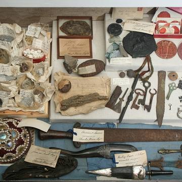 An overhead view of a tightly packed assemblage of approximately fifty objects. Most appear made of stone, metal, wax, or animal parts. Items include keys, weapons, wax seals, and a leather shoe. 