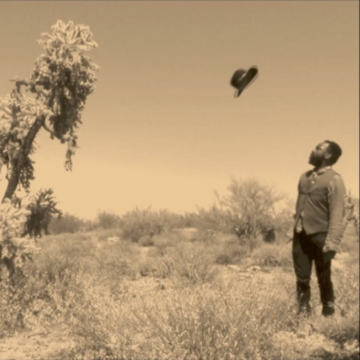 Sepia still from digital video Moses (Burning Bush), image depicting Samson Kambalu standing watching a hat thrown up in the air. The setting is in an arid looking landscape with shrubs and a small tree on the left.