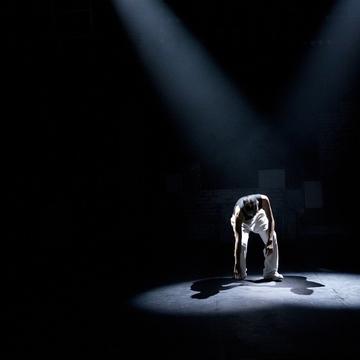 Image of performer touching one hand on the ground in spotlight on a dark stage