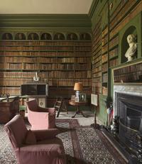 An image of a heavily book-lined room, with classical busts ornamenting the shelves. Two chairs sit in front of the fireplace.