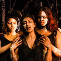 two women of colour hold and look at Medea (middle)who laments. White tribal markings on faces, wearing plain black clothing. Trees are in the background. 