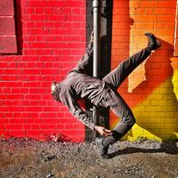 Man in black clothes, bending backwards with his left foot in the air, holding on to a drainpipe attached to a red, orange and yellow wall 